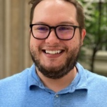 A headshot of Brody DeBettignies. He has short brown hair, a beard, and dark-rimmed glasses. He's wearing a blue polo shirt in an outdoor setting.