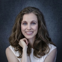 A headshot of Kristin Marrs. She has medium length brown hair and she's wearing a white sleeveless top in front of a blue-gray background.