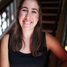 A headshot of Meredith Healy. She has brown hair, a big smile and is wearing a black tanktop in front of a staircase.