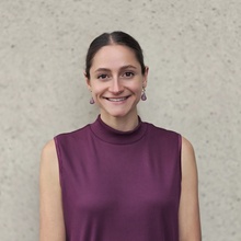 A shot of Sarah Frank in front of a neutral colored wall. She's wearing a raspberry-colored tanktop with her hair pulled back.