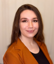 A professional headshot of Hailey McCoy-Munger on a tan background. She has brown hair and an dark orange blazer.