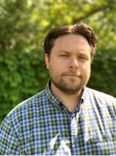 A headshot of Jake Tappen against a background of green leaves. He has a beard and is wearing a blue and green plaid shirt.